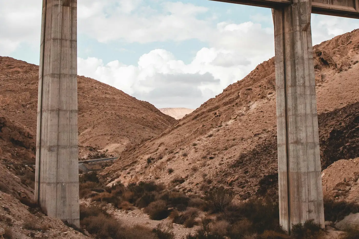 Concrete freeway overpass pillars rising above a dry desert canyon with rocky hills