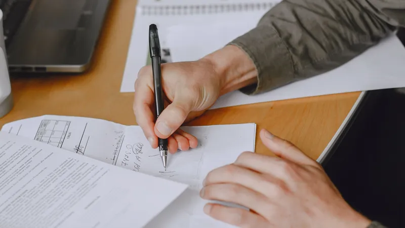 Close-up of a hand writing with a black pen on paperwork at a wooden desk beside a laptop