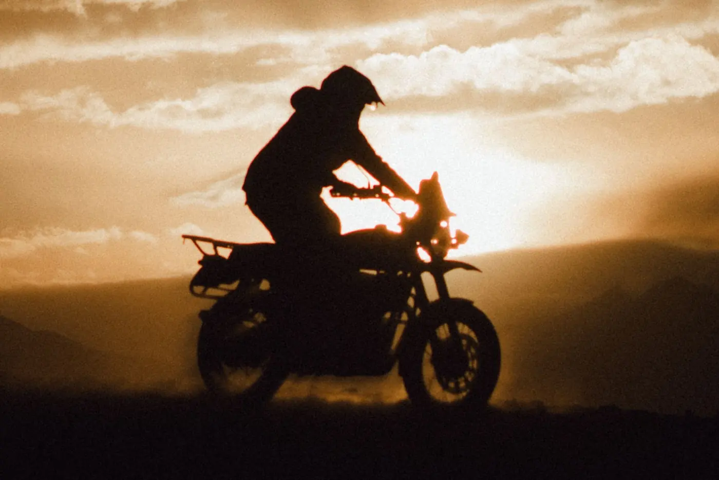 Silhouetted motorcyclist riding across dusty ground at sunset with hazy mountains in the background